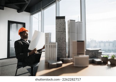 Office interior with panoramic city view. Handsome chief architect man in business suit and protective helmet holding blueprint sitting at desk working with city buildings model.