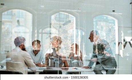 Office Life. Group Of Business People Discussing Something While Sitting At The Office Table Behind The Glass Wall In The Modern Office. Meeting