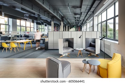 Office In A Loft Style With Large Windows, Gray Walls And Concrete Columns. There Are Many Workplaces With Computers And Shelves And Lockers, Relax Zone With Armchairs With Round Tables And Lamps.