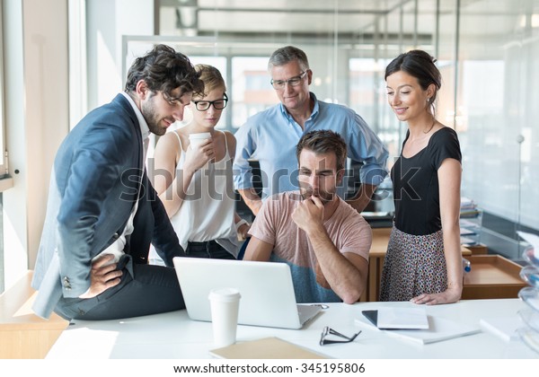 Office meeting, everybody is looking at the\
computer engineer website presentation. He is sitting at a desk in\
a luminous open space, the team is standing around him with the\
grey hair senior partner