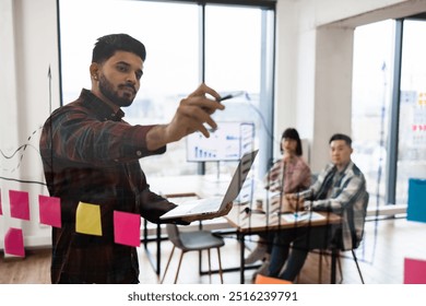 Office meeting featuring brainstorming session with colleagues. Man writing on glass wall with sticky notes and graphs in background. Team collaboration and business strategy planning in workspace. - Powered by Shutterstock