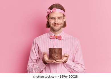 Office Party Concept. Cheerful Man Celebrates Anniversary At Workplace Receives Congratulations From Colleagues Holds Small Cake With Burning Candle Wears Big Spectacles And Festive Clothing.