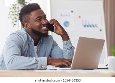 Office Phone Etiquette. Cheerful African American Employee Talking On Cellphone And Using Laptop At Workplace.