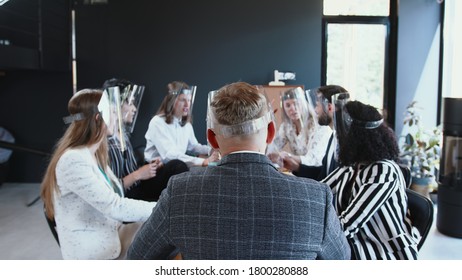 Office Safety Measures During COVID-19. Rear View Male Boss And Diverse Team Wear Face Shields At Business Meeting Table