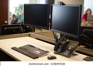 Office Setting - Dual Monitor Computer And Phone On Desk With Working Adults Out Of Focus In Background