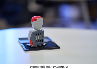 Office Stamp with Four Rolling Wheels, Resting on an Open Stamp Pad Filled with Blue Ink. Red Details on the Stamp Head and Body. White, Curved Office Desk. Blurred Background, Window Reflections