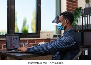 Office Startup Employee With Covid Mask Typing On Laptop Working On Market Research Pie Chart. Entrepreneur Wearing True Wireless Earbuds Using Portable Computer For Analyzing Business Sales Data.