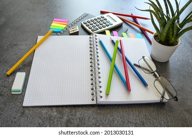 Office Or Study Desk With An Open Spiral Notebook, Colored Pencils, Calculator And Stationary Supplies On A Dark Gray Table, Copy Space, Selected Focus, Narrow Depth Of Field