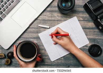 Office table desk, Flat lay. Workspace with laptop, red cup, candy, lens and flash on wooden background. Woman hand writing in a notebook