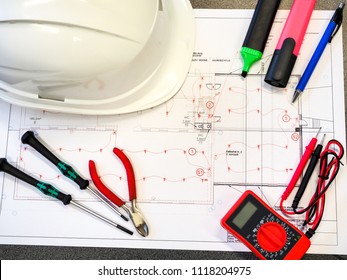 Office Table Or Desk With The Project Drawings Of The Security Or Fire Alarm System, A Protective Helmet, Tools, A Multimeter And Markers.