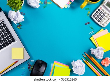 Office table desk with set of colorful supplies, white blank note pad, cup, pen, pc, crumpled paper, flower on blue background. Top view and copy space for text. - Powered by Shutterstock