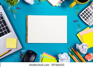 Office Table Desk With Set Of Colorful Supplies, White Blank Note Pad, Cup, Pen, Pc, Crumpled Paper, Flower On Blue Background. Top View And Copy Space For Text