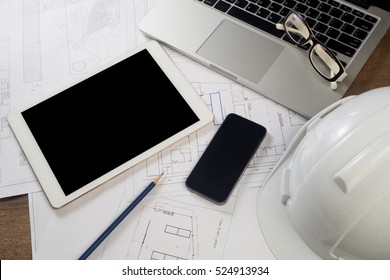 Office Table With Tablet, Cell Phone On Layout Paper. Engineering Office Place With Communication Object And Anti Knock Helmet On Wood Table. 