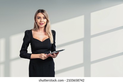 Office Woman In Black Suit Smiling, Looking At The Camera, Digital Device In Hands. Office Company And Staff. Copy Space Blank Wall. Concept Of Secretary In Business Company