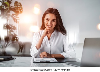 Office Woman Happy Look In White Office Room, Bokeh Lights, Laptop And Clipboard With Papers On The Table. Desk With Notebook And Technology. Concept Of Business Secretary