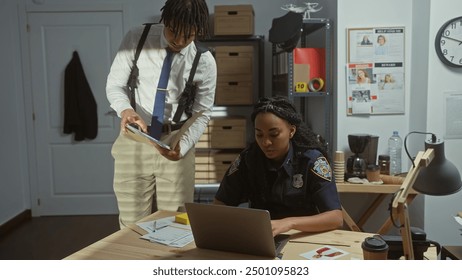 In an office, a woman officer examines her laptop while a man consults notes, both strategizing an investigation. - Powered by Shutterstock