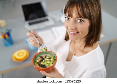 Office Woman Sitting At Her Desk, Smiles At The Camera While Holding Her Healthy Meal In A Bowl.