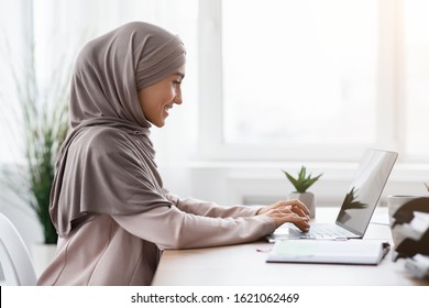 Office Work For Islamic Women. Happy Muslim Girl In Hijab Working On Laptop Computer At Desk, Typing On Keyboard, Side View With Free Space