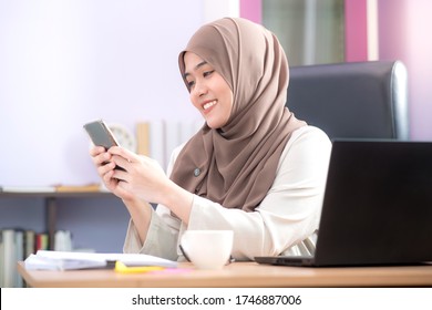 The office worker, an Asian Muslim woman, is sitting in front of her laptop computer at her desk and playing mobile phones and smiling happily at the office. - Powered by Shutterstock