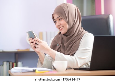 The office worker, an Asian Muslim woman, is sitting in front of her laptop computer at her desk and playing mobile phones and smiling happily at the office.