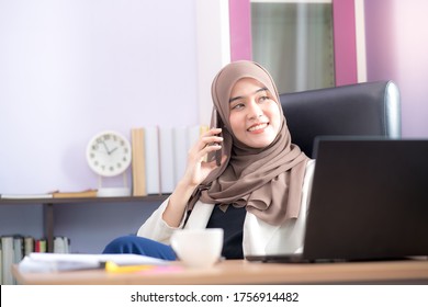 An office worker for an Asian Muslim woman sitting in front of a laptop computer at her desk and talking on a mobile phone and working at the office. - Shutterstock ID 1756914482