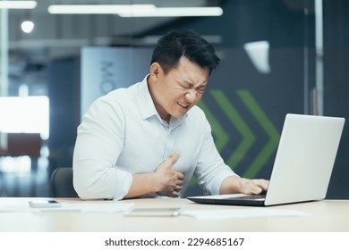 An office worker of Asian origin is working on a laptop and holding his chest with his hand. Suffering and writhing in pain, he needs medical help. - Shutterstock ID 2294685167