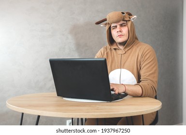 Office Worker In Cosplay Costume Of A Cow. Guy In The Funny Animal Pyjamas Sleepwear Near The Laptop. Man Is Working From Home. Search Job, Unemployment  Concept, Economy Crisis. Remote Work.