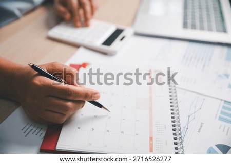 Office worker marking calendar while calculating financial data, illustrating schedule planning, time management, business tasks, productivity, professional workflow in modern workplace environment.