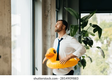 Office worker standing by window, wearing a duck swimming ring and imagining a relaxing beach holiday