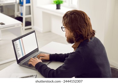 Office worker using a laptop computer. Male manager or financial accountant working on a modern notebook PC. Busy man sitting at his desk and typing on the keyboard. Back view, over the shoulder