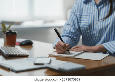 Office worker is writing notes in notebook at desk in modern office, keyboard, mouse, smartphone, calculator, documents and coffee are visible on the desk