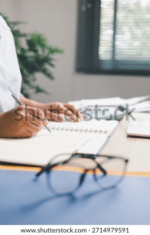 Office worker writing notes in notebook while reviewing documents on desk, illustrating planning, organization, productivity, business tasks and professional workflow in modern workplace setting.