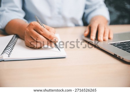Office worker writing notes in a notebook while using a laptop, illustrating planning, productivity, business tasks and professional workflow in modern workplace environment.