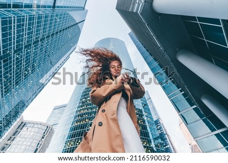 Office worker young woman wearing fashion clothes waits for business meeting looking at clock and holds laptop in arms standing against financial center. 