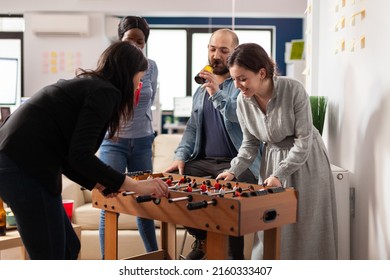 Office workmates playing foosball table game together after hours, enjoying alcoholic drinks and pizza. Man and women doing party celebration and having fun with match after work.
