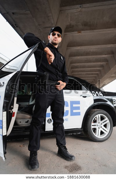 Officer of police in sunglasses and uniform\
standing near car on urban\
street