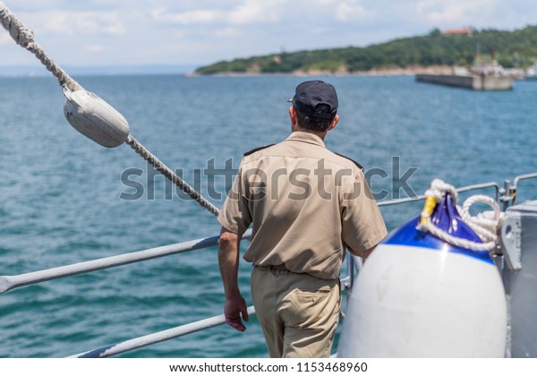 Officer in uniform on the deck of a warship. a\
soldier of the naval force goes on the deck of a battleship and\
looks at the harbor in the\
distance.