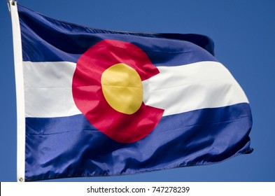 The Official Flag Of The State Of Colorado Waves Boisterously Against A Brilliant Blue Sky.  This State Sits In The Middle Of The United States, With The Rocky Mountains Running Through Its Center.