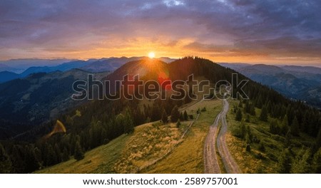 Offroad path crossing hill top. Dark dirt road illuminated with bright setting sun. Amazing sunset scenery in wild mountain forest. Beautiful nature landscape