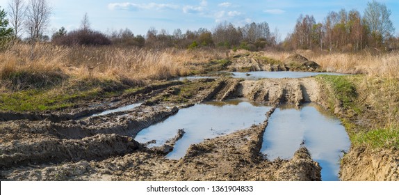 Off-road track. Tire imprints. Puddle detail. Military area. Impassable way. Muddy bumpy path in spring panoramic terrain. Bare trees, green bushes and dry grass in deserted rural landscape. Blue sky.