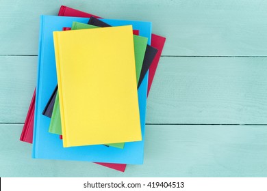 Offset Stack Of Multicolored Hardcover Books Piled Up On A Stained Pastel Colored Green Wood Table With A Bright Yellow Book On Top And Copy Space To The Side, Overhead View