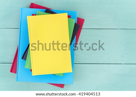 Offset stack of multicolored hardcover books piled up on a stained pastel colored green wood table with a bright yellow book on top and copy space to the side, overhead view