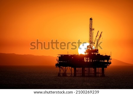 Offshore oil platform off the coast of California frames against an orange sky full of smoke from a nearby fire as the sun sets behind the rig.

