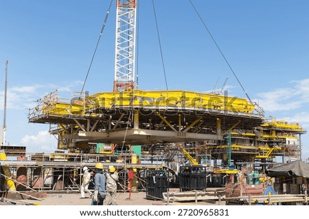 Offshore platform under construction lifted by towering crane. Steel structure, mechanical systems installed at oil gas energy site. Represents heavy industry, engineering and energy sector investment