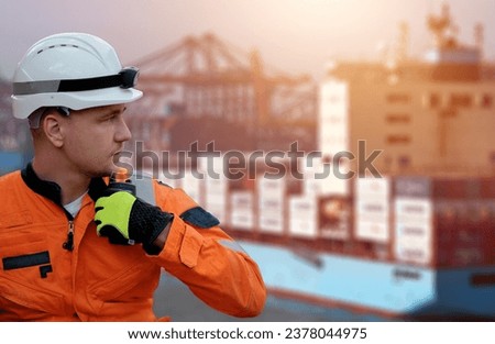 Offshore technician. Seafarer. Seaman. Navigator. A man in a boiler suit wearing a safety helmet with a container ship in the background. Seafarer in front of the container ship with a radio station.