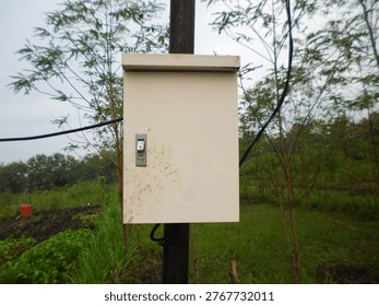 An off-white electrical control box with a switch mounted on a wooden pole in a lush green outdoor environment, surrounded by trees and vegetation under a cloudy sky.