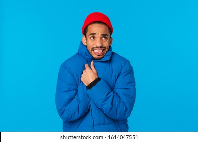 Oh gosh its freezing cold. Young man walking street feeling discomfort, trembling from low winter temperature, wrap himself, cover body in padded jacket and red hipster beanie, chattering teeth