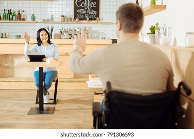 Oh Hi. Well-built Blond Disabled Man Sitting At The Table In A Cafe And Waving To A Beautiful Cheerful Dark-haired Woman Sitting Opposite Him