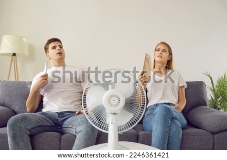 Oh, it's so hot today. Our home is overheated. Young couple exhausted from summer heat. Two tired sweaty people use both electric and paper fans to make some fresh air in sweltering summertime weather