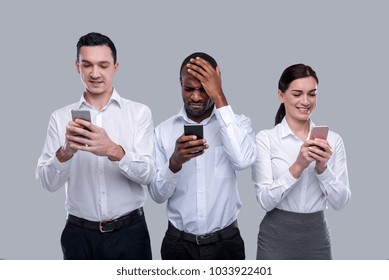 Oh Man. Handsome Well-built Afro-american Young Man Touching His Forehead And Using His Phone While His Colleagues Standing Near His And Typing On Their Phones